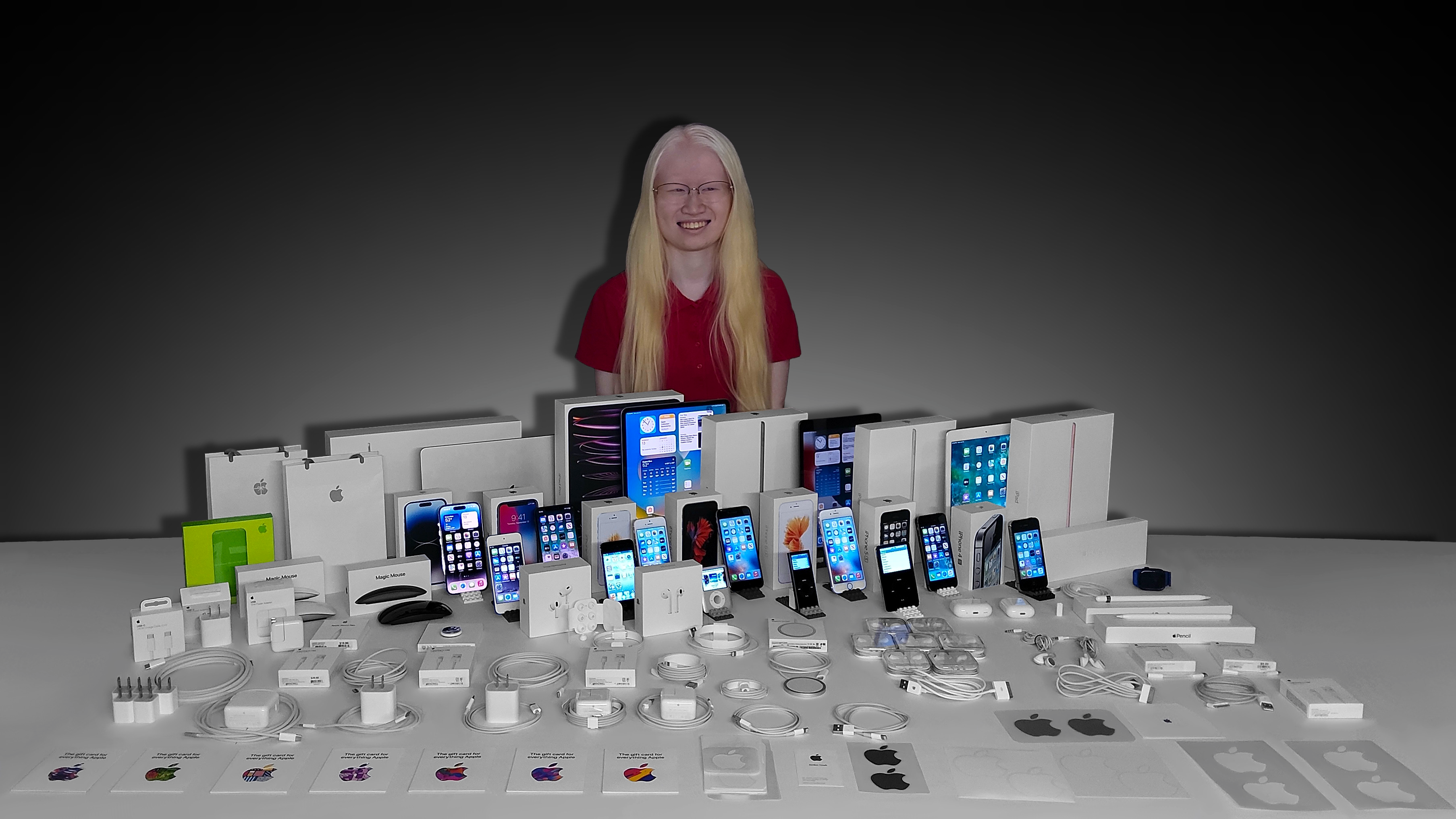 A picture of Leah Dykema kneeling on the floor by a desk with her Apple collection arranged to be displayed. She wears a t-shirt with symbals of Apple products across it.