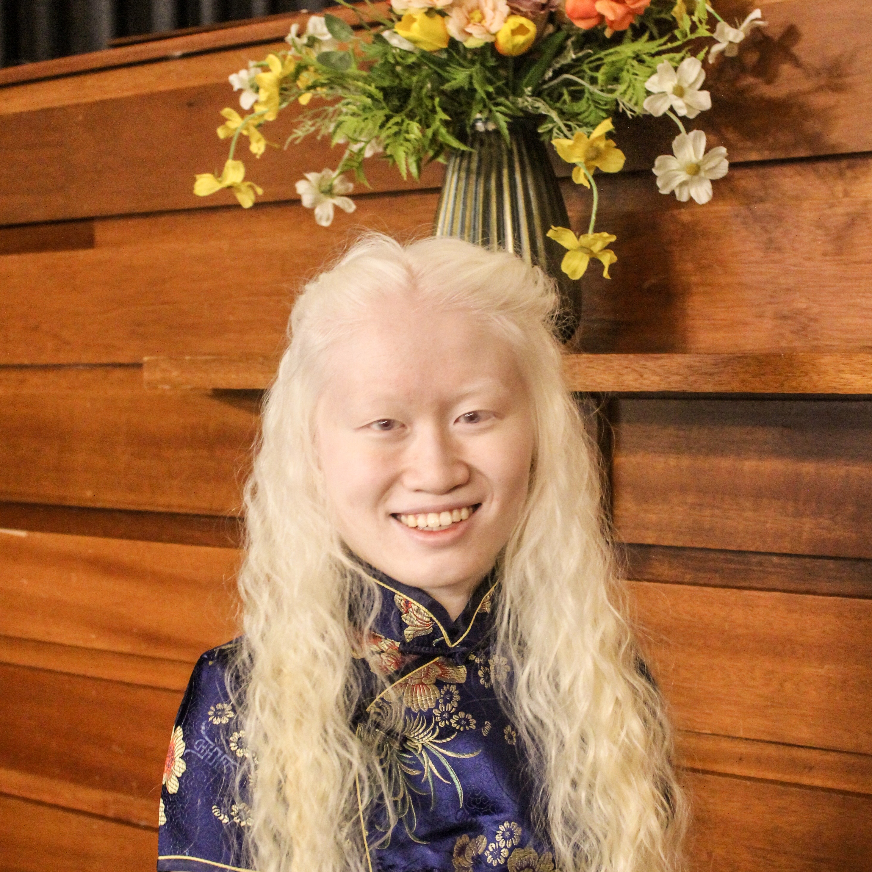 A close up picture of Leah Dykema as she smiles at the camera with simple glasses and a bun with braids in her hair. She wears a red Chinese cheongsam dress with ornate gold design.
