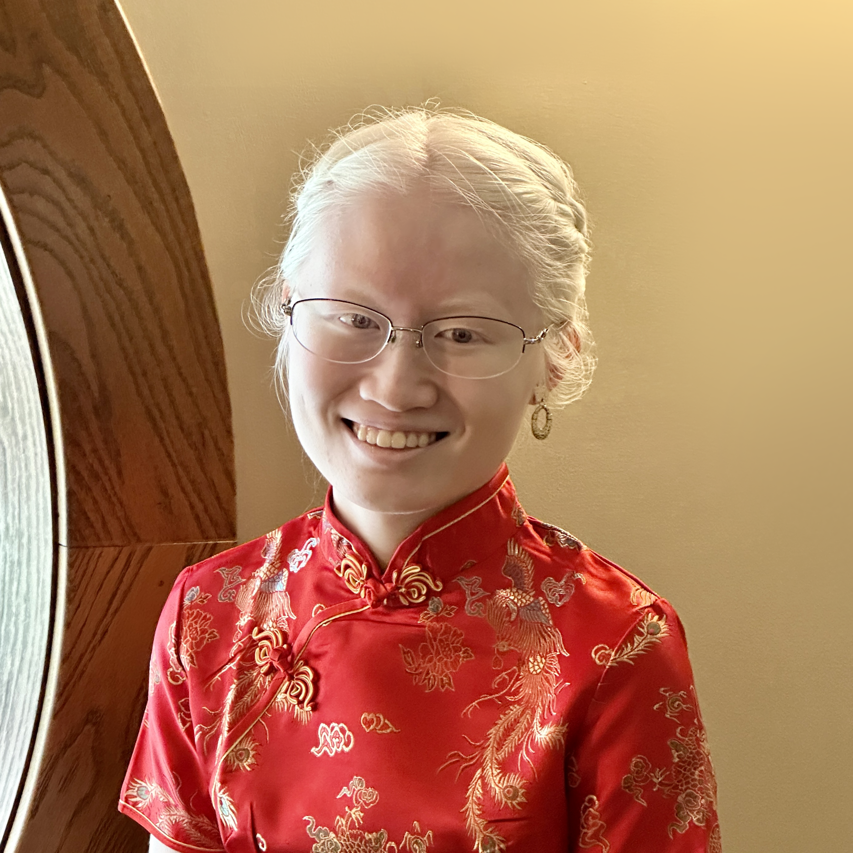 A close up picture of Leah Dykema as she smiles at the camera with simple glasses and a bun with braids in her hair. She wears a red Chinese cheongsam dress with ornate gold design.